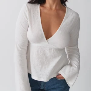 Vit omlottopp med knyt  - Supersnygg vit omlottopp med djup v-ringning och långa ärmar. Toppen har en lätt strukturerad yta och knytband i midjan som ger en fin siluett. Perfekt att styla med jeans eller kjol för en trendig look.