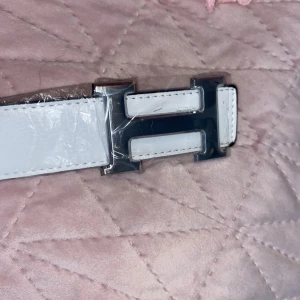 Vitt skärp med H-spänne - Snyggt vitt bälte med ett stort, silvrigt H-format spänne. Bältet har stilrena sömmar längs kanterna och är gjort i slätt skinn. Perfekt accessoar för att lyfta din outfit med en clean och modern look. Storlek 110cm