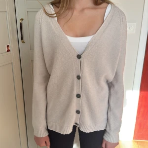 Beige kofta med knappar och v-ringning - Mysig beige kofta med djup v-ringning och fyra stora knappar framtill. Långärmad modell med ribbade muddar och nederkant. Perfekt att slänga över ett linne eller t-shirt för en chill och stilren look.