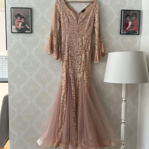Champagnefärgad långklänning med spets - Säljer en elegant långklänning i champagnefärg från Mireck. Klänningen har v-ringning, utsvängda långärmar och är täckt av glittriga paljetter och blommig spets. Nederdelen är i mjuk tyll som ger en snygg volym. Perfekt för bal eller nyår!