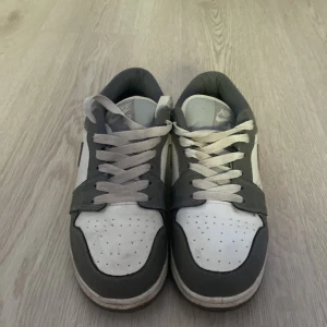 Nike Dunk Low i grått och vitt - Snygga Nike Dunk Low sneakers i en clean färgkombo av grått och vitt. Skorna har klassisk siluett med rund tå, perforerad tåbox och vit sula. Ovandelen är i skinn med gråa overlays och vit bas, samt matchande grå swoosh och snörning.
