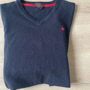 Mörkgrå ulltröja från Morris - Snygg mörkgrå stickad tröja från Morris i 100% ull. V-ringad modell med lång ärm och röd broderad logga på bröstet. Perfekt för dig som gillar klassisk stil med en twist av färgdetalj vid halsen. (PRIS KAN DISKUTERAS) storlek s/m