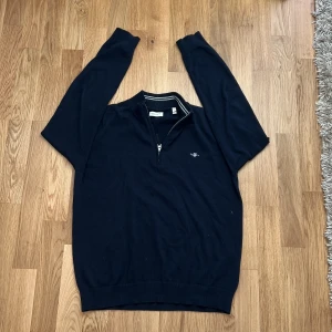 Mörkblå half zip tröja från Gant - Snygg mörkblå långärmad tröja från Gant med half zip och liten broderad logga på bröstet. Tröjan har ribbade muddar och krage med diskreta ränder på insidan. Perfekt för dig som gillar klassisk och stilren look.