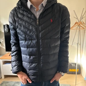 Marinblå dunjacka från Ralph Lauren - Säljer en svart dunjacka från Ralph Lauren med huva och dragkedja. Jackan har quiltad design och den klassiska röda loggan på bröstet. Perfekt för kyliga dagar och riktigt snygg till jeans eller chinos.