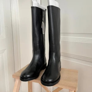 Svarta läderstövlar från Zara - Stilrena svarta stövlar från Zara i läder med dragkedja på insidan. Modellen är knähög med platt sula och rund tå, perfekt till höstens outfits. Snygg minimalistisk look som funkar till många stilar.