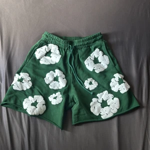 Denim Tears shorts - Gröna shorts från Denim Tears med elastisk midja och snörning. De har unika vita blommiga tryck över hela plagget och en bakficka. Sköna och loose fit, perfekta för sommaren. Materialet är mjuk bomull som känns chill och avslappnat.