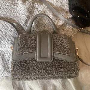 Grå axelväska med teddy-detaljer - Säljer en snygg grå axelväska med teddy-liknande material och detaljer i syntetläder. Väskan har ett handtag upptill och en lång justerbar axelrem. Perfekt för att lyfta din outfit med en trendig touch!