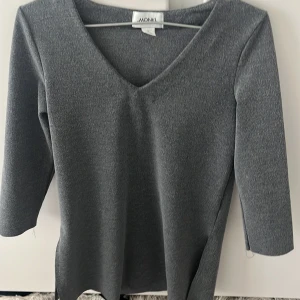 Grå klänning med v-ringning från Monki - Säljer en stilren grå klänning från Monki med v-ringad hals och trekvartsärm. Klänningen har en rak passform och är tillverkad i ett mjukt material som känns bekvämt mot huden. Perfekt för dig som gillar enkel och tidlös stil.
