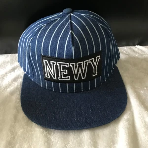 Blå keps med vita ränder och NEWY - Snygg blå keps med vita vertikala ränder och stort broderat NEWY-märke framtill. Klassisk snapback-modell med justerbar passform. Perfekt för en cool och avslappnad stil.