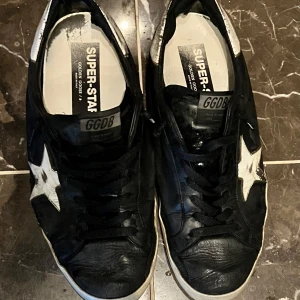 Svarta Golden Goose med vit stjärna - Säljer ett par svarta Golden Goose sneakers med vit stjärndetalj på sidan och vit sula. Skorna har snörning och rund tå, samt märkets logga bak på hälen. Klassisk och trendig modell med slitna detaljer. 100% äkta bevis och box, behöver pengar för pirots 4 som är incoming, är såklart öppen för förhandlingar 
