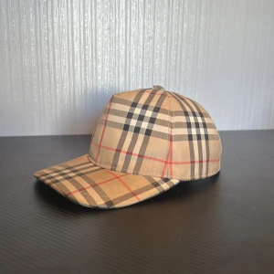 Beurberry keps - Säljer en klassisk keps från Burberry i beige med det ikoniska rutmönstret i svart, rött och vitt. Kepsen har justerbar snapback baktill och en böjd skärm. Märke med 'Burberry London England' bak.