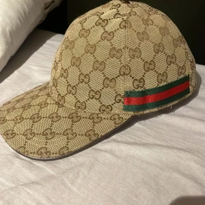 Beige keps från Gucci  - Säljer en beige keps från Gucci med klassiskt GG-monogram över hela kepsen. Kepsen har grönt och rött band på sidan och justerbart spänne bak. Perfekt för dig som gillar lyxiga accessoarer och vill sticka ut.