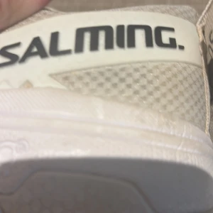 Vita sneakers från Salming - Säljer ett par vita sneakers från Salming med ljusgrå detaljer och mönstrad ovandel. Skorna har snörning och en gummisula i brunt med hexagonmönster undertill. Perfekta för dig som gillar sportig stil. Kontakta gärna vid frågor, pris:140kr