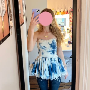 Blåvit tubtop med fransar - Supersnygg axelbandslös top i denim tyg. Toppen har markerad midja, volangig nederdel och fransig kant med en dragkedja på baksidan. Den har inga defekter och är bara använd en gång. Säljer eftersom jag rensar ut kläder jag inte använder längre. 