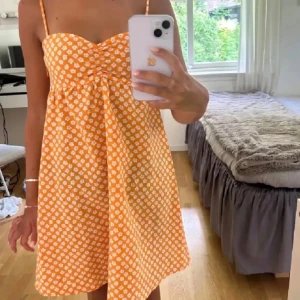 Orange blommig sommarklänning med smala axelband - Säljer en söt orange klänning med vita blommor och smala axelband. Klänningen har en lätt utställd passform och är perfekt för varma dagar. Den är kort och har en feminin, somrig känsla.