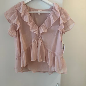Ljusrosa volangblus - Söt ljusrosa blus med volanger längs ärmar, halsringning och nederkant. Blusen har korta ärmar och v-ringning med knyt framtill. Perfekt för en romantisk och feminin stil. Helt ny med prislapp kvar🥰