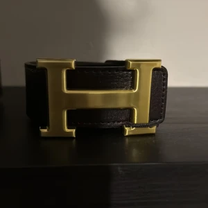Brunt skärp med guldfärgat H-spänne - Säljer ett brunt skärp från Hermès med ett stort H-format spänne i guldfärg. Bältet är gjort i skinn och har en stilren, klassisk design som passar till många olika outfits.