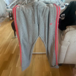 Grå mjukisbyxor från Adidas med rosa detaljer - Grå mjukisbyxor från Adidas med rosa ränder längs sidorna och rosa Adidas-logga på låret. Byxorna har dragsko i midjan och raka ben. Perfekta för en sportig och avslappnad stil.