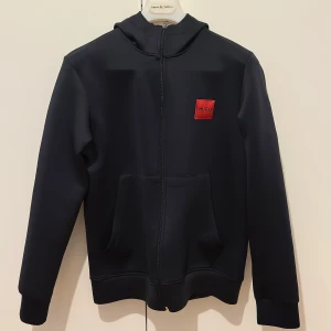 Svart hoodie från Hugo Boss - Säljer en svart hoodie från Hugo Boss med dragkedja och huva. Tröjan har en röd logotyp på bröstet och praktiska fickor framtill. Pris :249kr