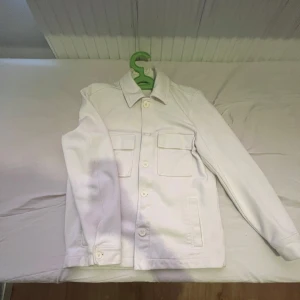 Vit overshirt med knappar - Säljer en stilren vit overshirt med klassisk krage och stora bröstfickor. Jackan har knäppning framtill och raka ärmar. Perfekt för lager-på-lager och enkel att matcha med olika outfits.