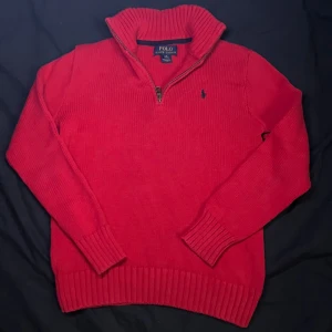 Röd stickad tröja med dragkedja från Polo Ralph Lauren - Säljer en röd stickad tröja från Polo Ralph Lauren med halv dragkedja och hög krage. Tröjan har långa ärmar, ribbade muddar och en liten blå logga broderad på bröstet. Perfekt för kyliga dagar.