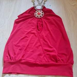 Röd halterneck topp med smyckesdetalj - Snygg röd halterneck topp med dekorativ smyckesdetalj i halsen. Kedjor. Och som en stor brosch. Tillverkad i mjuk och stretchig polyesterblandning. Perfekt för dig som vill sticka ut med en färgstark och unik topp. Perfekt till fest, party. Använd en gång. Den passar xs-s