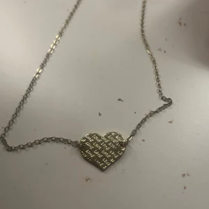 Halsband med hjärta i guldfärg - Säljer ett fint halsband med en tunn kedja och ett hjärtformat hänge i guldfärg. Hänget har ingraverad text som ger en personlig touch. Perfekt att bära till vardags eller ge bort som present.