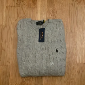 Grå kabelstickad tröja från Polo Ralph Lauren - Säljer nu en klassisk grå kabelstickad tröja från Polo Ralph Lauren. Tröjan har rund halsringning, lång ärm och den ikoniska broderade loggan på bröstet. Perfekt för lager-på-lager och stilrena outfits. Det är storlek S men pssar större! Pris kan diskuteras.
