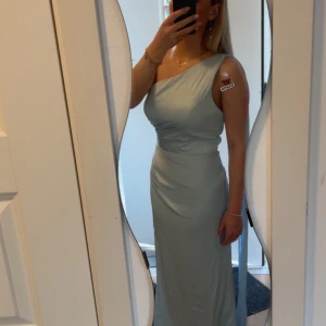 Balklänning  - Säljer en jättefin ljusblå balklänning långklänning med oneshoulder och draperad midja. Klänningen har en öppen rygg och ett stilrent, figurnära snitt. Helt oanvänd då den inte passar mig (min kompis på bild) sista bilden är från hemsidan fast i annan färg 