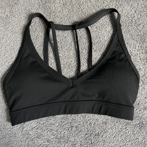 Svart sport bh - Säljer en svart sport bh med smala dubbla axelband och v-ringning. Toppen har en stilren design och är perfekt för träning eller som ett snyggt basplagg. Materialet känns mjukt och stretchigt.