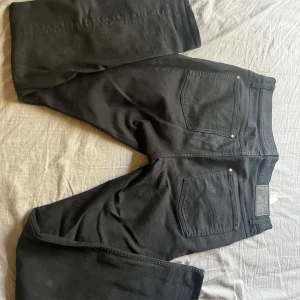 Svarta jeansbyxor från Tiger of Sweden - Säljer ett par svarta jeans från Tiger of Sweden. Klassisk femficksmodell med raka ben och diskreta detaljer. Jeansen har svarta sömmar och en svart läderpatch bak i midjan.