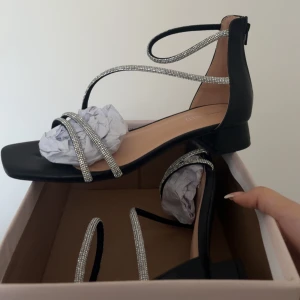 Svarta sandaler med glittriga remmar - Säljer ett par svarta sandaler med fyrkantig tå och glittriga remmar som går över foten och runt vristen. Skorna har låg klack och dragkedja bak. Perfekta för att lyfta vilken outfit som helst!
