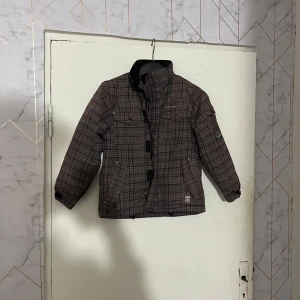 Brun rutig vinterjacka från Coaster Clothes - Säljer en brun rutig vinterjacka från Coaster Clothes i barnstorlek 122. Jackan har hög krage, dragkedja och knappar framtill samt två fickor. Perfekt för kalla dagar och har ett klassiskt rutigt mönster.