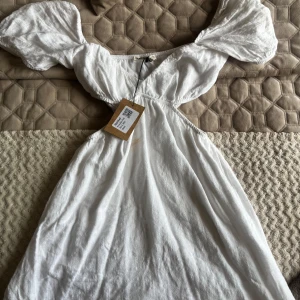 Vit klänning med puffärmar från Pull & Bear - Superfin vit studentklänning från Pull & Bear med puffiga kortärmar och markerad midja. Klänningen har en lätt v-ringning och är sydd i ett tunt, luftigt material som passar perfekt till varma dagar. (Har en sminkfläck i mitten som troligtvis går att tvätta bort smidigt i tvätten)