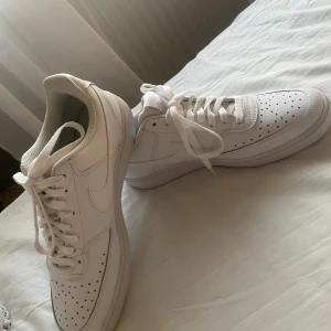 Nike Air Force 1 vita sneakers - Säljer ett par klassiska vita Nike Air Force 1 sneakers med perforerad tå och snörning. Skorna har en låg profil, rund tå och är tillverkade i skinn med vit sula. Skostorlek 43