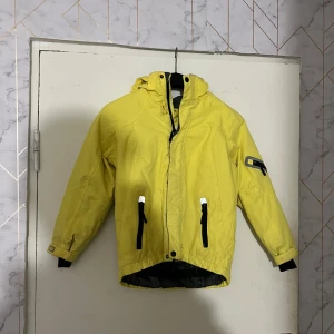 Gul vindjacka med huva - Snygg gul vindjacka med huva och svarta detaljer. Jackan har dragkedja framtill, två fickor med dragkedja och en extra ficka på ärmen. Perfekt för blåsiga dagar och enkel att matcha med streetwear.