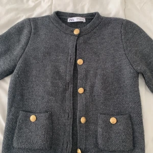 Grå cardigan jacka från Zara - Mysig grå cardigan jacka från Zara med rund halsringning och guldiga knappar framtill. Två fickor framtill med dekorativa knappar. Stickad i mjukt material, perfekt för lager-på-lager. Klassisk och stilren design.