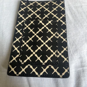Svart och beige mobilhållare  från By Malene Birger - Säljer en snygg korthållare från By Malene Birger i svart och beige med grafiskt mönster. Perfekt att lägga in mobilen och kort. Klassisk och stilren design med By Malene Birger-logga i guld. Man kan även lägga kort framför. Den är helt oanvänd 