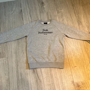 Grå sweatshirt från Peak Performance - Säljer en klassisk grå sweatshirt från Peak Performance med svart logga på bröstet. Tröjan har rund hals, långa ärmar och ribbade muddar vid ärmslut och nederkant. Perfekt för en avslappnad stil.Använd ett fåtal gånger.