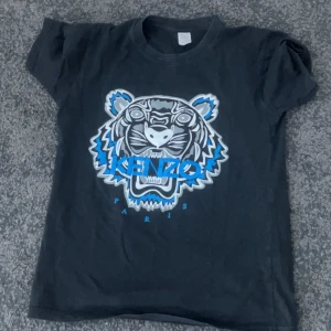 Svart t-shirt med tigertryck från Kenzo - Cool svart t-shirt från Kenzo med stort tigertryck i vitt, blått och grått på framsidan. Klassisk passform med rund hals och korta ärmar. Perfekt statement-plagg för dig som gillar streetwear och snygga loggor.