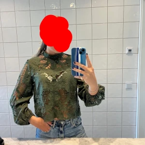 Mörkgrön spetsblus  - Säljer en grön blus med vackra spetsdetaljer och blommigt mönster. Blusen är croppad och har långa volangärmar samt hög hals