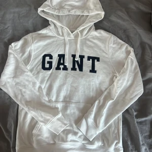 Vit hoodie från GANT - Säljer en klassisk vit hoodie från GANT med svart logotyp på bröstet. Tröjan har huva med dragsko och en stor magficka. Perfekt för en avslappnad stil. Är helt oanvänd och väldigt fint skick🌟