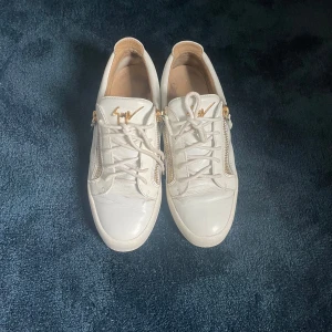 Vita sneakers från Giuseppe Zanotti med gulddetaljer - Säljer ett par vita sneakers från Giuseppe Zanotti med guldfärgade dragkedjor och logga. Skick 7/10 med tanke på creaasing annrs är dom i bra skick. Skriv till mig vid minsta fundering!