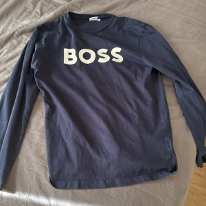 Mörkblå långärmad tröja från BOSS - Säljer en mörkblå långärmad tröja från BOSS med vit logotyp på bröstet. Tröjan har en klassisk rund halsringning och är tillverkad i mjuk bomull. Perfekt för en stilren och avslappnad look.