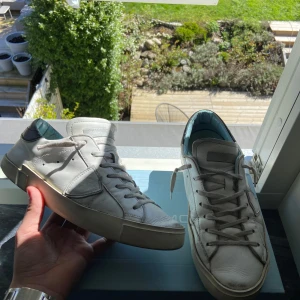 Vita sneakers från Axel Arigato - Säljer ett par vita sneakers från Philippe model med klassisk design och diskreta detaljer. Skorna har snörning, rund tå och platt sula. Insidan är ljusblå och ovandelen är i skinn. Perfekta för en stilren vardagslook.