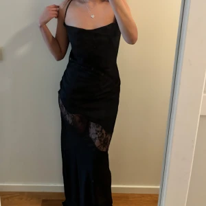 Svart långklänning med spets och öppen rygg - Säljer en svart långklänning med smala axelband och snygg öppen rygg med knyt. Klänningen har en transparent spetsdetalj över låren som ger en unik look. Perfekt för dig som vill sticka ut på festen! Köpt från meshki!