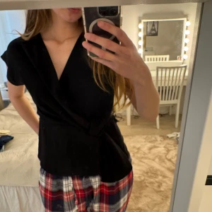 Svart omlottblus med bälte - Svart omlottblus med korta ärmar och knytbälte i midjan. Djup v-ringning och mjukt, följsamt material. Perfekt för en stilren look.