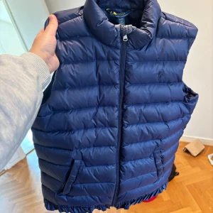 Ralph Lauren väst  - Säljer en blå dunväst från Polo Ralph Lauren med dragkedja framtill och två fickor. Västen har en hög krage och klassisk quiltad design, perfekt för lager-på-lager-stil.
