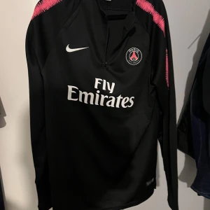 Svart Paris Saint-Germain fotbollströja med rosa detaljer - Säljer en svart långärmad PSG-tröja från Nike med rosa detaljer på axlarna och vitt Fly Emirates-tryck på bröstet. PSG-logga och Nike-märke framtill. Perfekt för dig som älskar fotboll och vill ha en snygg supportertröja.