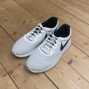 Vita Nike Tanjuns - Säljer ett par fräscha vita Nike sneakers med svart swoosh på sidan. Skorna har mesh-överdel för bra andningsförmåga, vita snören och en platt vit sula. 
